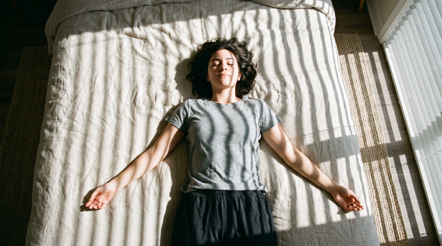 A woman lying on her back on a sunlit bed with her arms spread, relaxed